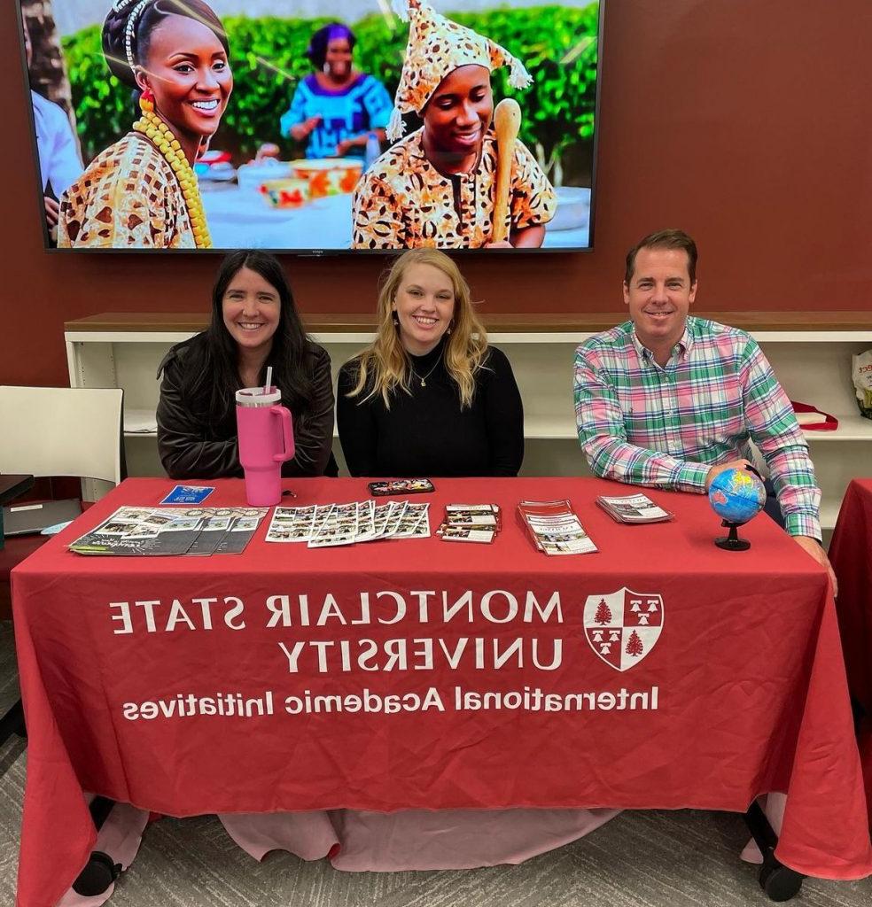 Group of three members of the Office of International Academic Initiatives in front of television with image of Burkina Faso cultural clothing. There is a man on the left side and two women to his right sitting in front of a table with a red tablecloth that has the office logo. 
